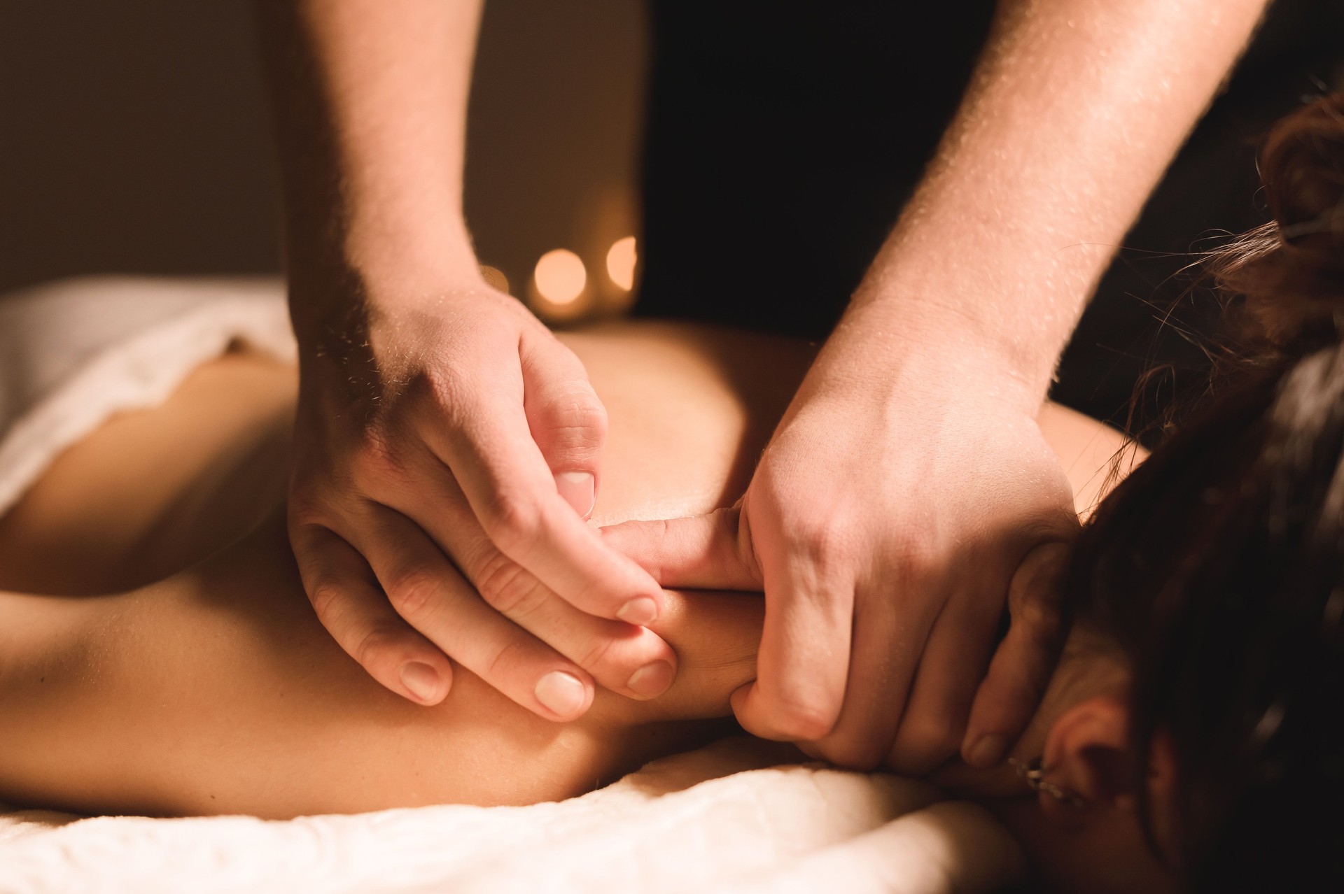 Men's hands make a therapeutic neck massage for a girl lying on a massage couch in a massage spa with dark lighting. Close-up. Dark Key
